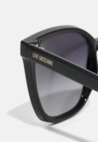 Μαύρα υπερμεγέθη γυαλιά ηλίου με gradient φακούς, που διαθέτουν γυαλιστερή επιφάνεια και χρυσή εκτύπωση "LOVE MOSCHINO" στον βραχίονα.