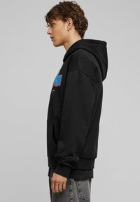 Zwarte hoodie met een voorzak, geribde manchetten en een kleurrijke grafische print op de borst. Zachte stof, casual pasvorm.