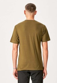 Mand med kort hår, iført en olivengrøn T-shirt med korte ærmer og sorte bukser, står med ryggen til mod en ensfarvet baggrund.