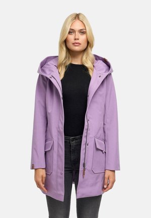 Blonde vrouw die een lichtpaarse capuchontrui draagt over een zwarte top en donkere spijkerbroek, kijkt recht vooruit met een neutrale uitdrukking.