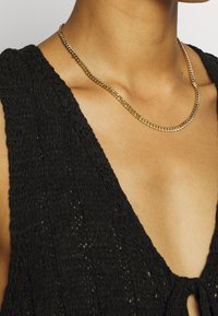 Gold-colored chain necklace with interlocking links, resting on bare skin, paired with a textured black knit top featuring a V-neck.