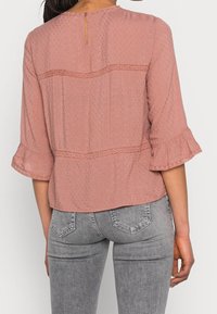 Blouse rose texturée avec manches trois-quarts à volants et ouverture en goutte d'eau au dos. Présente des accents en dentelle et une coupe ample.