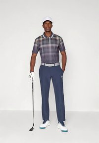 Vyras, dėvintis baltą kepuraitę, languotą golfo marškinėlį, tamsiai mėlynas kelnes ir baltus golfo batus, laiko golfo lazdelę ir stovi ant neutralios fono.