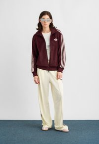 Mulher vestindo um casaco de zíper marrom da Adidas, calças largas creme, blusa branca, óculos de sol cinza, de pé sobre um carpete azul contra uma parede branca.