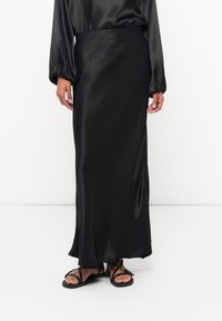 Fustă maxi din satin negru cu o siluetă fluidă, având o textură netedă și un ușor luciu, purtată cu sandale negre cu spirale.