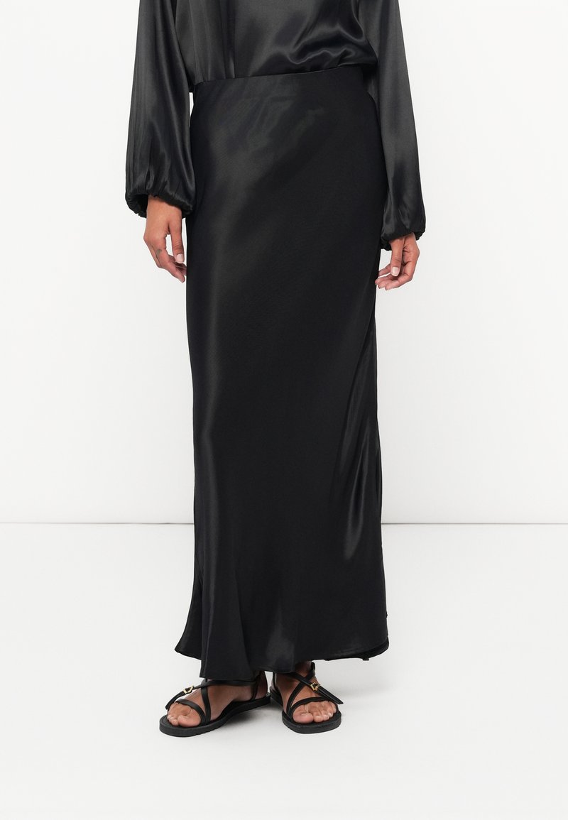 Sorte satin maxi-nederdel med en flydende silhuet, der har en glat tekstur og en let glans, båret med sorte spiral-sandaler.