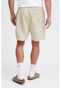 Beige shorts with an elastic waistband, single back pocket, and a relaxed fit, worn with white socks and brown slip-on sandals.
