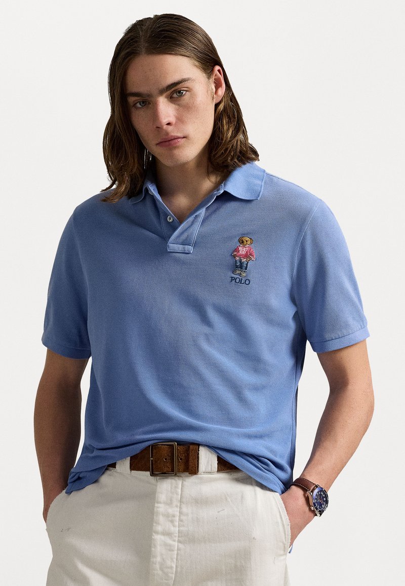 Light blue polo shirt made of textured fabric, featuring a bear graphic in a pink sweater and denim. Worn with white trousers and a brown belt.