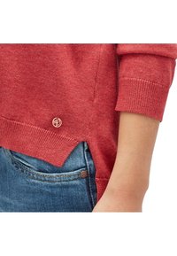 Röd tröja med ribbade ärmslut, med logotypdetalj vid fållen och en sidoklyv, bärs över blådenimjeans. Mjuk textur och lättviktsmaterial.