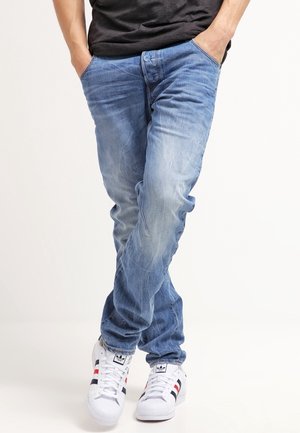 Persona con vaqueros azules desteñidos, camiseta negra y zapatillas Adidas blancas con rayas negras y rojas, de pie con las manos en los bolsillos.