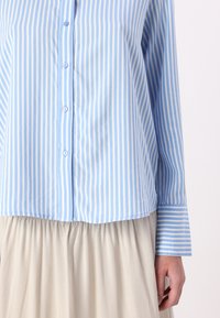 Light blue button-down shirt with white vertical stripes, featuring a rounded hem and long sleeves. Paired with a beige, gathered skirt.