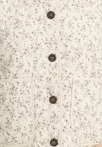 Prošívaná krémová látka s malým hnědým květinovým vzorem, vybavená čtyřmi tmavými kulatými knoflíky a dvěma předními našitými kapsami.