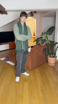 Zielony szalik na jasnozielonym swetrze, szare dżinsy i białe sneakersy; stojący na drewnianej podłodze w pokoju z rośliną i kanapą.
