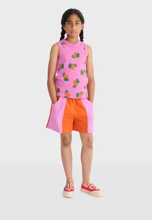 Djevojka stoji s rukama u džepovima, odjevena u ružičasti top bez rukava s printom naranče, narančasto-ružičaste kratke hlače i crvene cipele bez vezica.