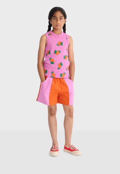 Fille debout, les mains dans les poches, portant un haut sans manches rose avec un motif de fruits orange, un short orange et rose, et des chaussures rouges sans lacets.