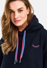 Mladá žena s dlouhými vlnitými vlasy, která má na sobě černou mikinu s růžovými a modrými šňůrkami a malou růžovou výšivkou řas na hrudi.