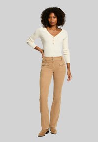 Suéter de punto beige claro con escote en V y tres botones, combinado con pantalones de pana anchos en color tostado que cuentan con bolsillos frontales y herrajes decorativos.