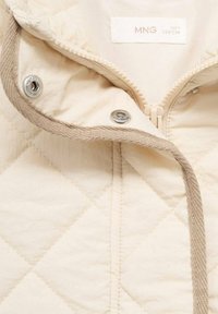 Veste matelassée beige avec fermeture éclair à l'avant et boutons-pression. L'intérieur présente une doublure en tissu doux. Accentuée d'une bordure beige et d'une surface texturée.