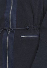 Chaqueta de forro polar azul marino con un acabado texturizado, que cuenta con una cremallera central, cintura con cordón y un bolsillo lateral con cremallera.
