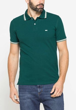 Uomo che indossa una polo verde scuro a maniche corte con colletto e bordi delle maniche a righe bianche, abbinata a jeans di denim blu.
