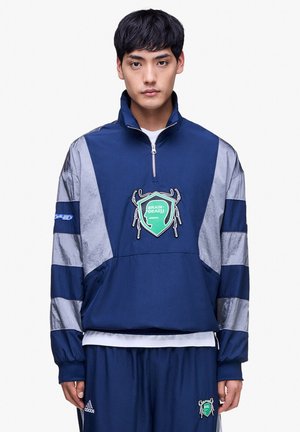 Joven con chaqueta deportiva azul marino y plateada con un emblema verde "Brain Dead" y pantalones Adidas azul marino a juego.