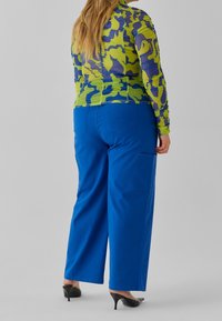 Maglietta a maniche lunghe con motivo astratto verde e blu, abbinata a pantaloni blu a vita alta con tasche laterali e design a gamba dritta.