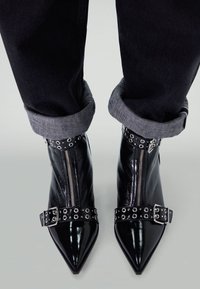 Black patent leather ankle boots with pointed toes, silver zippers, and studded straps. Worn with rolled-up dark jeans.