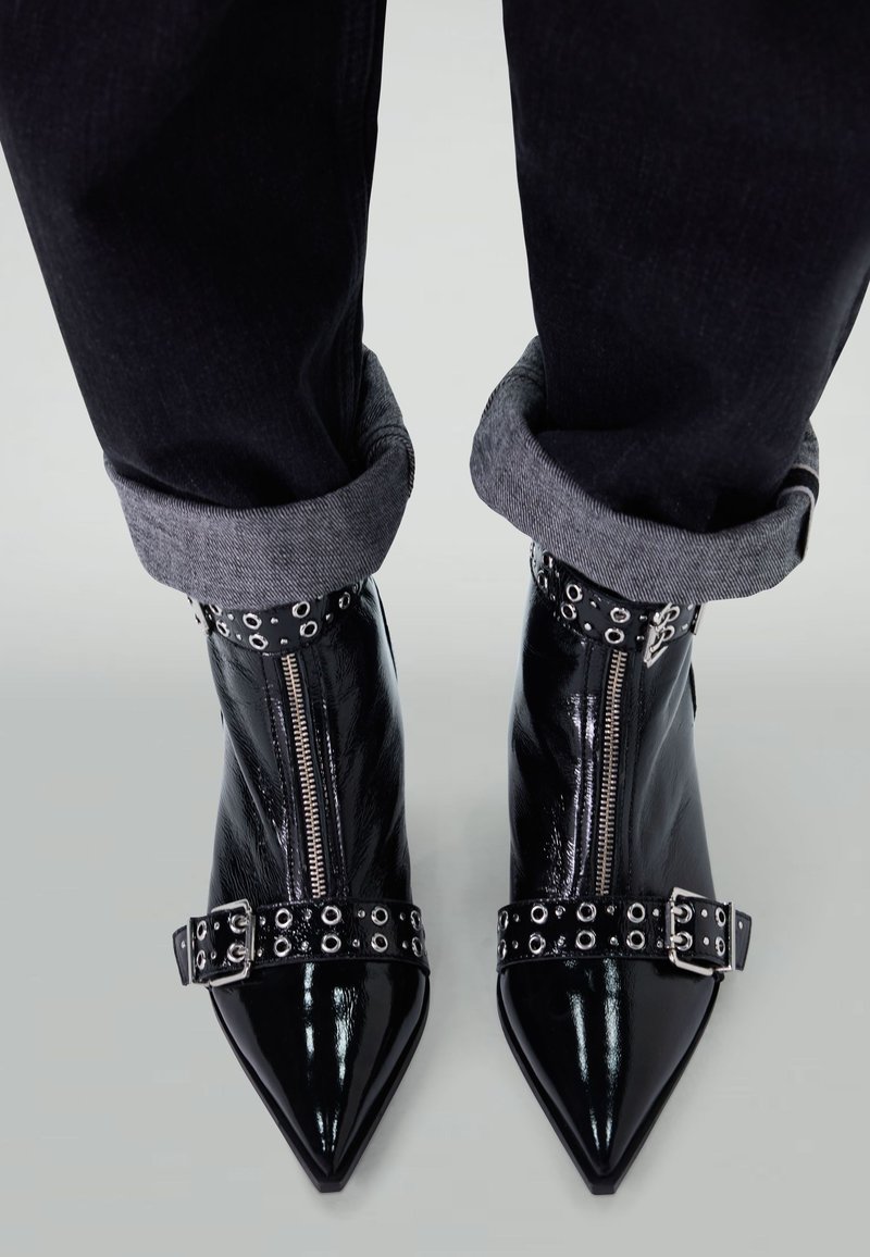 Black patent leather ankle boots with pointed toes, silver zippers, and studded straps. Worn with rolled-up dark jeans.