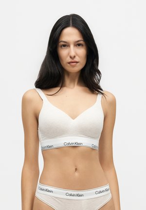 Femme aux longs cheveux foncés portant un soutien-gorge Calvin Klein beige clair et des sous-vêtements assortis sur un fond blanc uni.