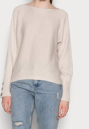 Femme portant un pull en maille crème avec des boutons aux manches et un jean bleu clair déchiré, debout devant un fond uni.