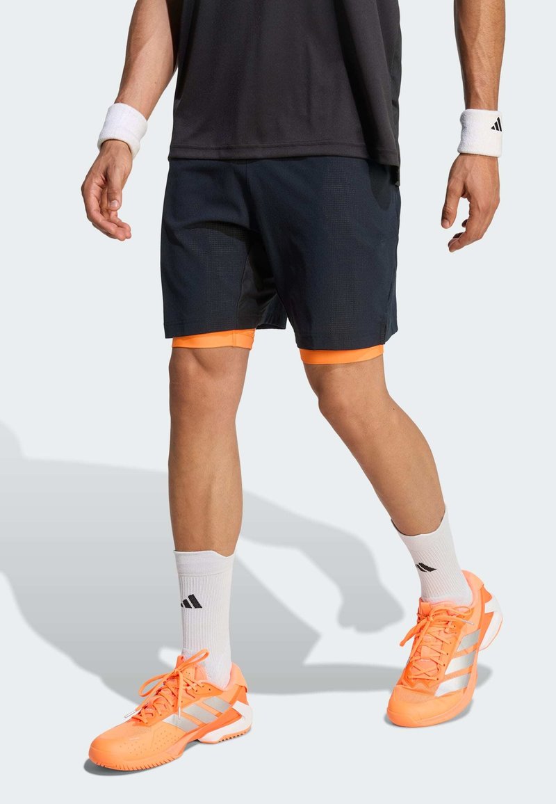Shorts de sport marine avec bordure orange, associés à des chaussures de sport orange vif avec des accents blancs. Chaussettes blanches avec détail logo noir.