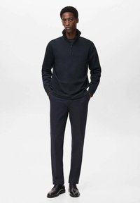 Sudadera negra con cremallera hecha de tejido de punto, combinada con pantalones oscuros y zapatos negros. El modelo está de pie con las manos en los bolsillos frente a un fondo claro.