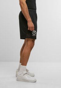 Male wearing black Colucci shorts, white socks, and white Nike Air Force 1 sneakers, standing against a plain light background.