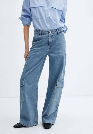 Personne portant une chemise bleu clair boutonnée et un jean taille haute à jambes larges avec des poches cargo, debout sur fond blanc.