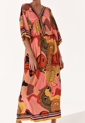 Femme portant une longue robe colorée à imprimé abstrait avec un décolleté en V et des manches larges, debout devant un fond uni.