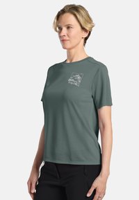 Chemise à manches courtes en tissu vert foncé avec un col rond et un graphique de montagne sur le côté gauche de la poitrine. Texture lisse, coupe décontractée.