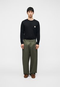 Μαύρο πουκάμισο με μακριά μανίκια και τσέπη στο στήθος, συνδυασμένο με φαρδιά πράσινα παντελόνια και καφέ παπούτσια, στέκεται μπροστά σε λευκό φόν το.