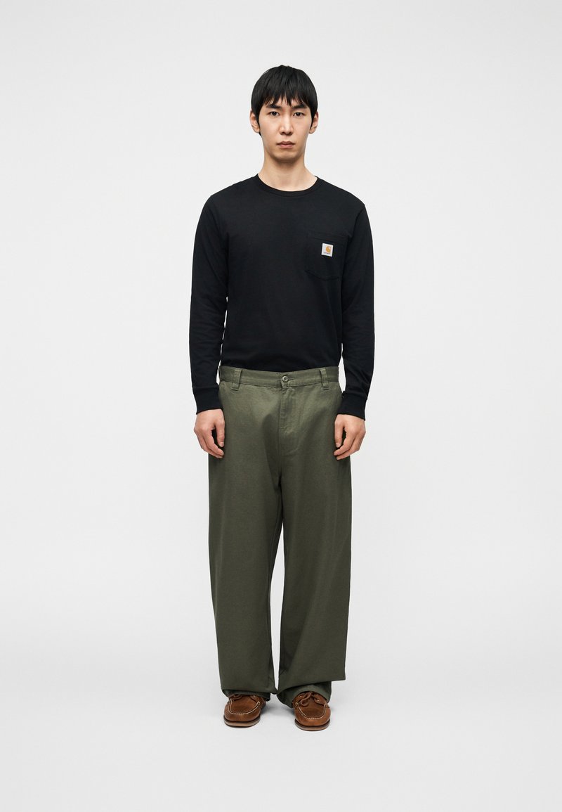 Μαύρο πουκάμισο με μακριά μανίκια και τσέπη στο στήθος, συνδυασμένο με φαρδιά πράσινα παντελόνια και καφέ παπούτσια, στέκεται μπροστά σε λευκό φόντο.