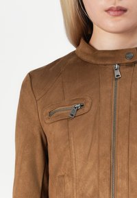 Veste en suède marron avec un col montant, fermeture éclair à l'avant et poche zippée diagonale. Elle présente des coutures ton sur ton et une texture lisse.