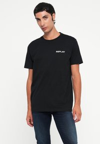Sort bomulds t-shirt med rund hals og korte ærmer. Forsiden viser "REPLAY" i hvid tekst på venstre side.
