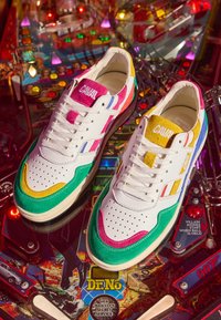 Paire de baskets en cuir blanc avec des touches vertes, roses, jaunes et bleues sur une surface colorée de machine à flipper.
