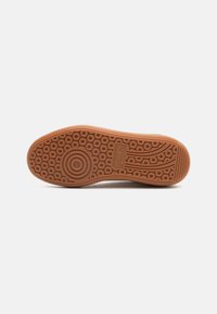 Suela de zapato en goma marrón claro con patrones de tracción circulares y ondulados. Textura suave con un ligero brillo, presentando detalles del logo de la marca.