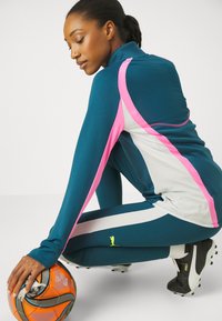 Teal treningstøy med rosa og hvite detaljer, lange ermer og en tettsittende design. Modellen sitter på kne ved siden av en oransje fotball, iført svarte fotballsko.