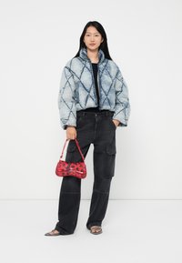 Vrouw in een blauwe gewatteerde jas, zwarte cargobroek, zwart top, met een kleine handtas met een rood rozenpatroon en zwarte sandaaltjes met straps.