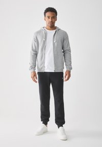 Mann steht vor einem schlichten Hintergrund und trägt einen grauen Reißverschluss-Hoodie, ein weißes T-Shirt, schwarze Jogginghosen und weiße Sneakers, und blickt nach vorne.