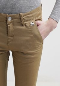 Slim-fit beige byxor i bomullsmaterial. Har en framficka, silverknappstängning och en liten logotypplik på sidan.