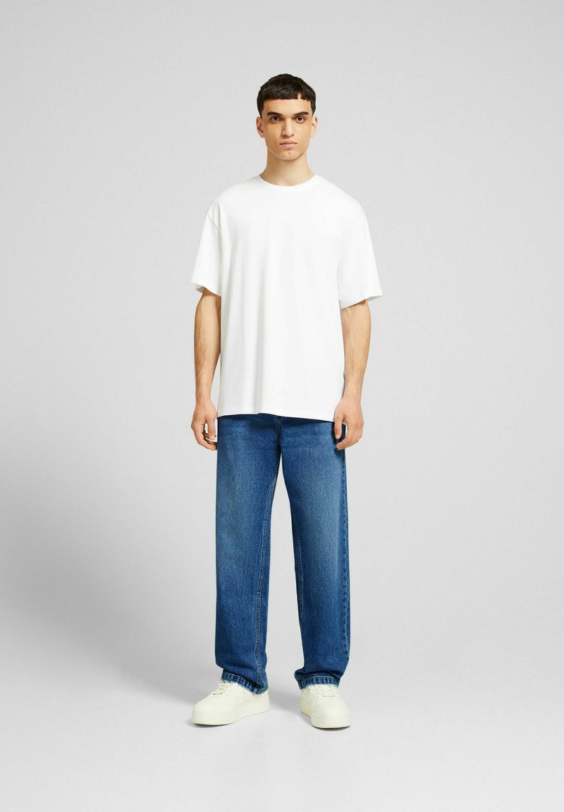Hombre vestido con una camiseta blanca básica de gran tamaño, jeans rectos azules y zapatillas blancas, de pie contra un fondo gris claro.