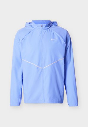 Veste coupe-vent Nike bleu clair avec fermeture éclair intégrale à l'avant, capuche, manches longues et bandes réfléchissantes blanches en forme de V sur le devant.