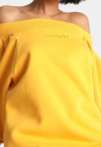 Sudadera amarilla con los hombros al descubierto, hecha de una tela suave. Presenta un cuello acanalado y un logotipo en relieve de "IVYPARK" en el frontal.