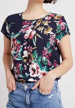 Kobieta ubrana w granatowy krótki rękaw z dużym kwiatowym nadrukiem i jasnoniebieskie dżinsy z wysokim stanem, z rękami w kieszeniach.